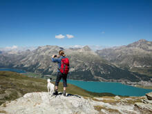 Eine Frau steht mit einem Hund auf einem Berg und macht mit ihrem Smartphone ein Bild von der Landschaft.