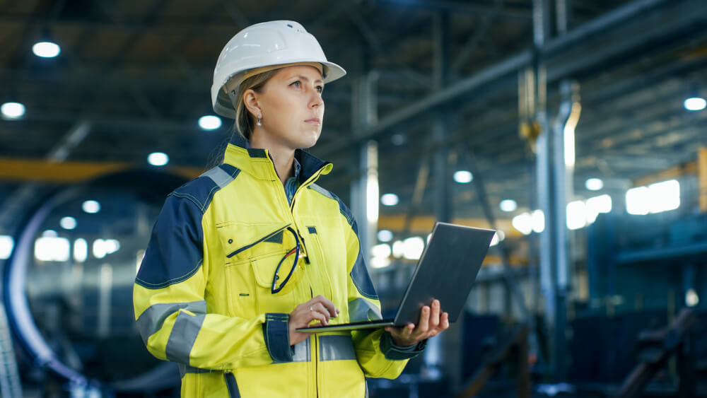 Eine junge Ingenieurin steht mit ihrem Laptop in einer Produktionshalle und analysiert die Vorgänge.