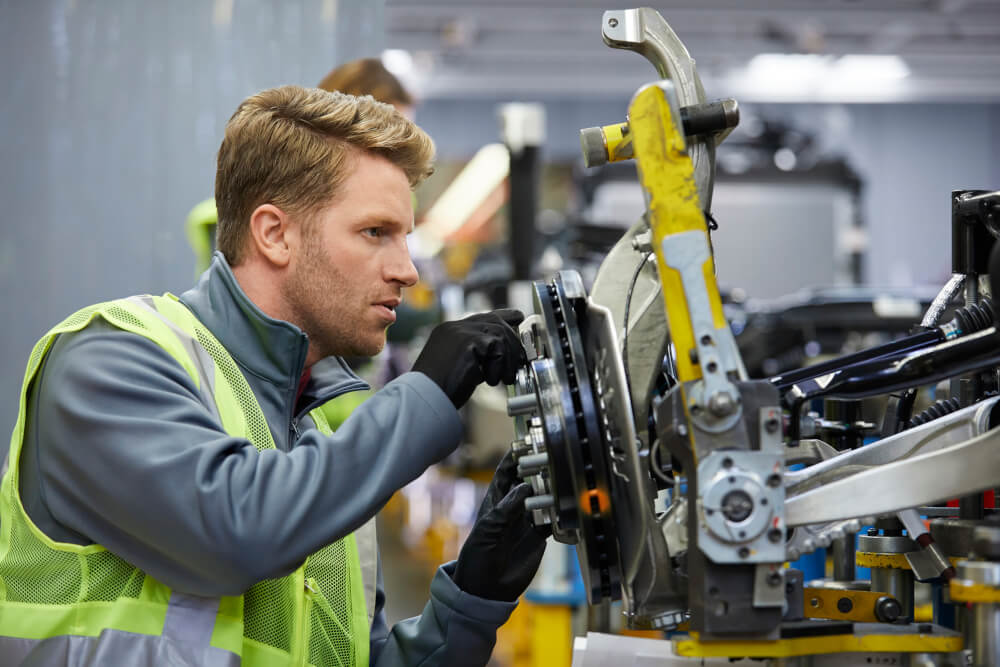Männlicher Ingenieur untersucht das Fahrgestell eines Autos in der Autoindustrie-Fabrik. Er trägt reflektierende Kleidung.