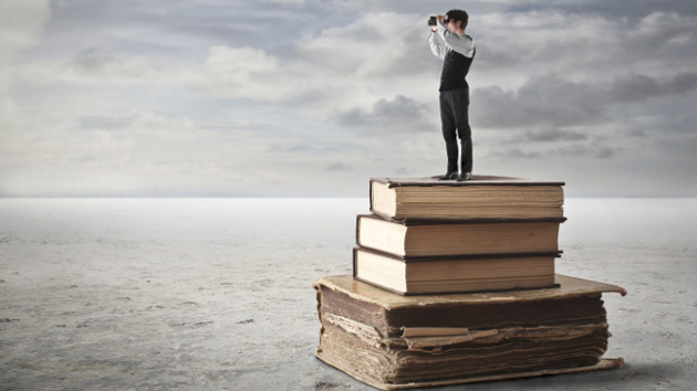 Eine kleine Person steht auf einem Stapel alter Bücher und schaut durch ein Fernglas in die Ferne.